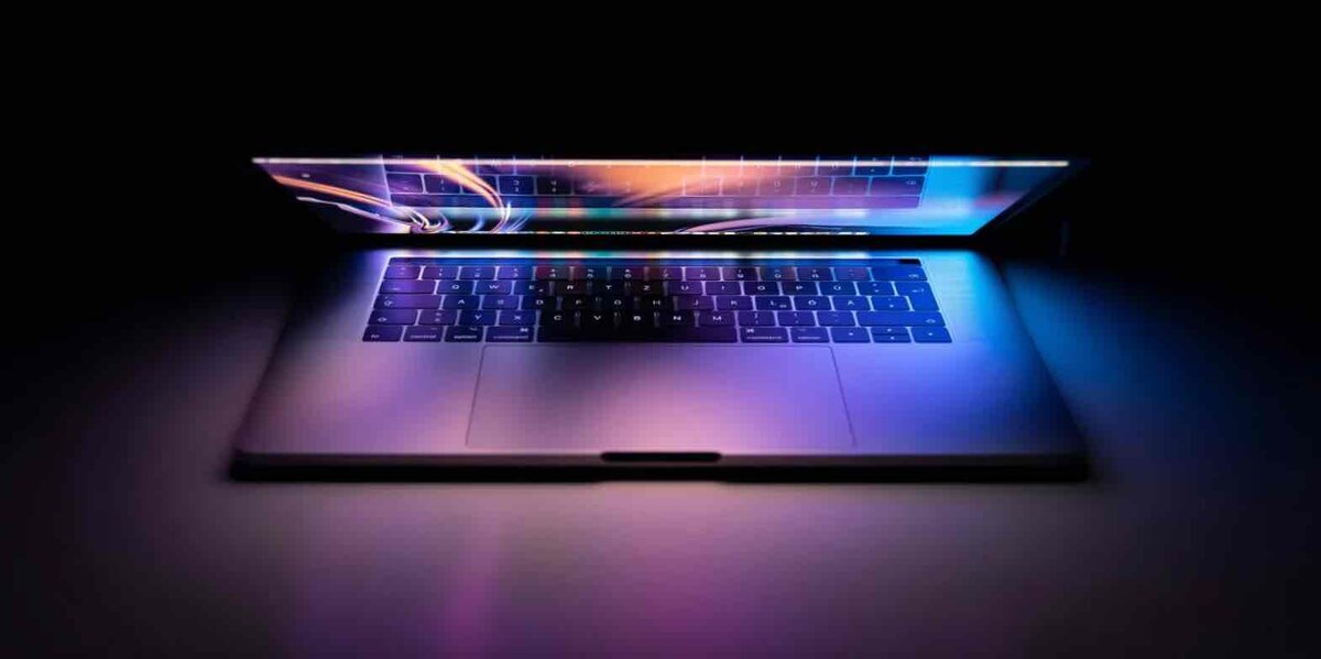 暗い背景の中で、半開きのMacBookの画面とキーボードに青や紫の光が反射している様子
