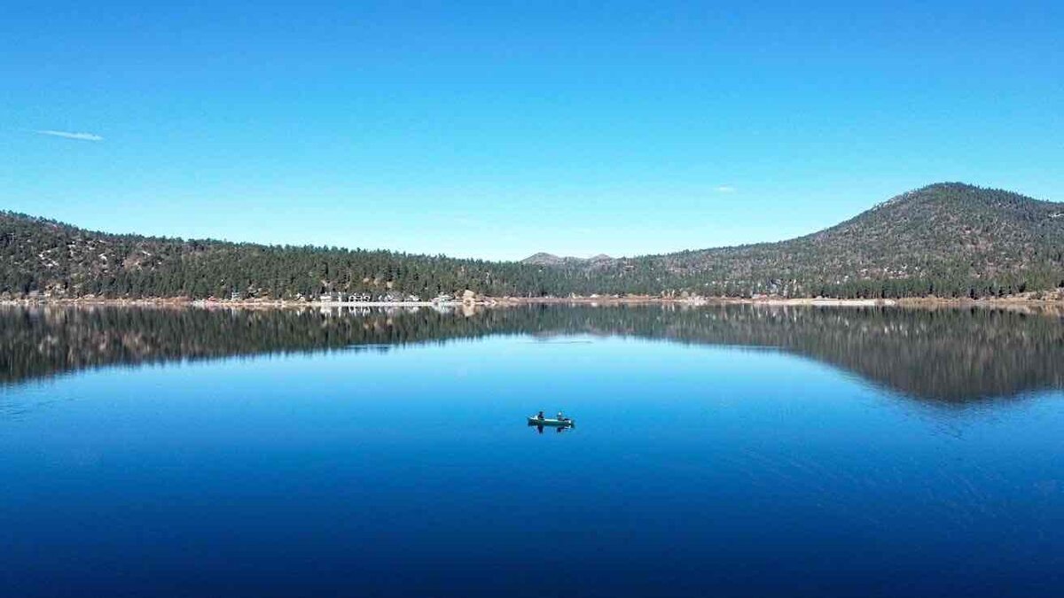 青空の下、森と山に囲まれた湖面に景色が反射し、中央に小さなボートが浮かんでいる風景