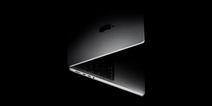 黒い背景にシルバーのノートパソコンを半分だけ開いた状態。天面にAppleロゴ、左側面にUSB-C型のポートが2つ見え、隙間からキーボードがのぞいている
