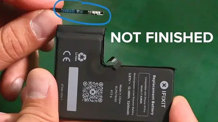 手に持たれたiPhone用の黒い交換バッテリー。iFIXITのロゴと「Compatible with iPhone 14 Pro」「Model # A2865」のラベルがあり、上端のコネクタ部分が青い丸で囲まれ、白字で「NOT FINISHED」と書かれている