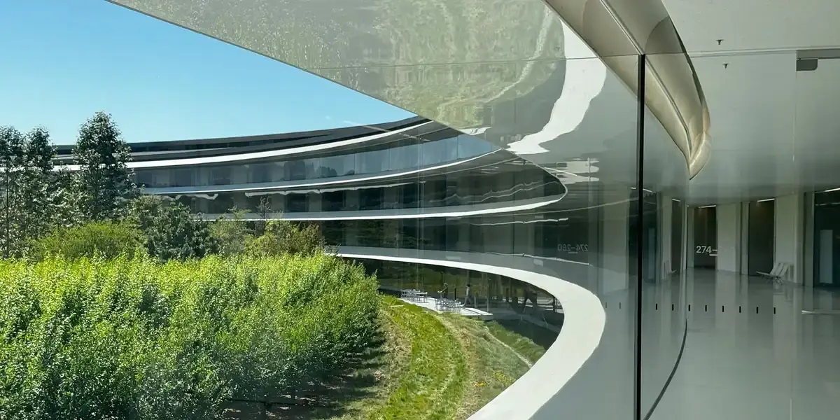 晴れた空の下、Apple Parkの円形の社屋。曲面ガラスの通路越しに中庭の緑と対岸の建物が映り込んでいる