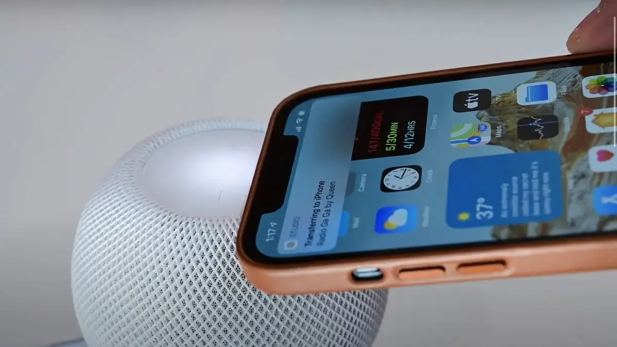 白いHomePod miniの上面すぐ近くにiPhoneを持っていったところ。iPhoneの画面上部に「Transferring to iPhone」と曲名を示す受け渡しのバナーが表示されている