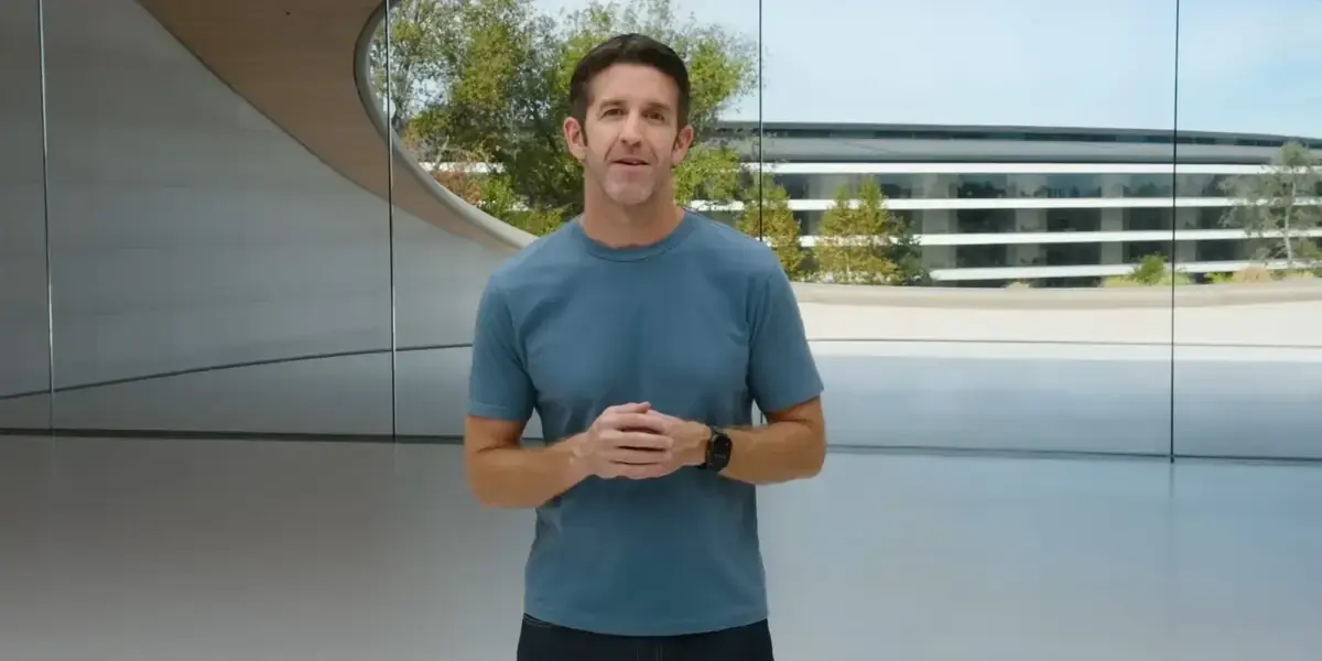 Apple Parkのガラス張りの建物を背に、青いTシャツ姿の男性が正面を向いて話している