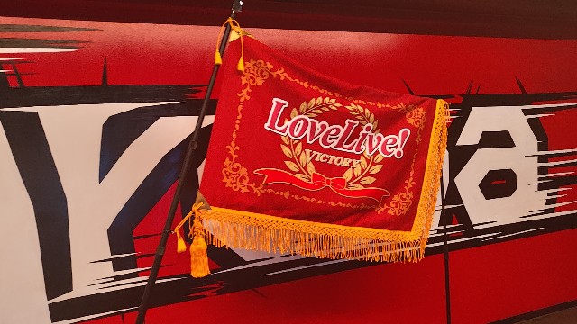 スクールアイドルの大会「ラブライブ」の優勝旗です。赤い布地に大会の紋章が刻みつけられており、雄大な姿です。蓮ノ空女学院スクールアイドルクラブ 102期卒業公演会場である、Kアリーナ会場内にて設置・展示されていました。