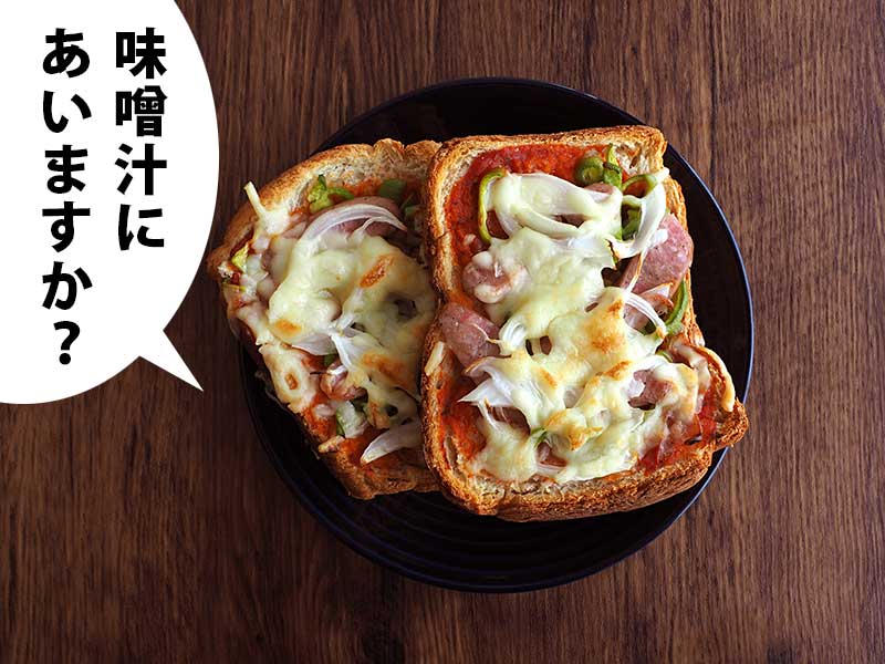 木の机にピザトーストを乗せた黒い皿が置かれている。吹き出しには「味噌汁にあいますか?」と書いてある。