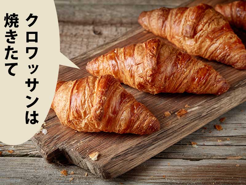 机の上のウッドボードに乗ったクロワッサン３つ。吹き出しに『クロワッサンは焼きたて』