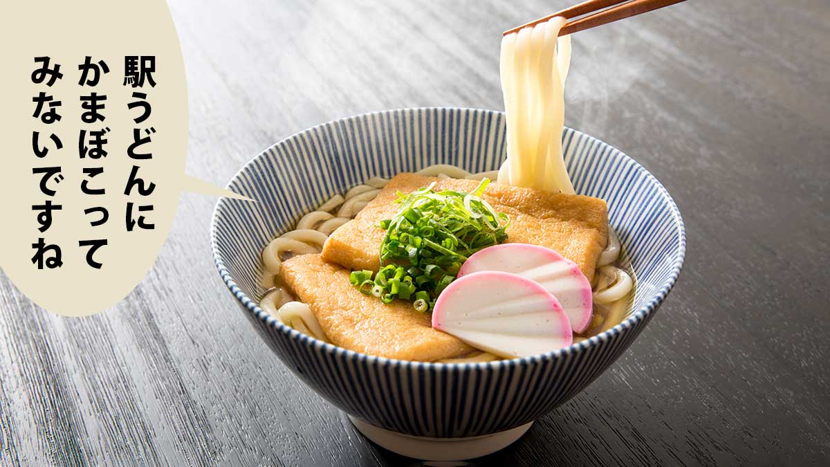 黒い机の上に乗ったきつねうどん。かまぼことネギがついていて、箸で麺をあげている。吹き出しに「駅うどんにかまぼこってみないですね」