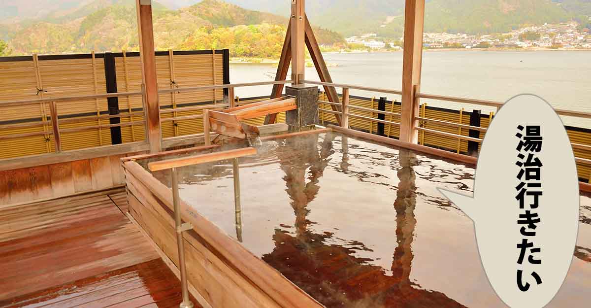 遠くに山と湖が見える木で出来た露天風呂。吹き出しに「湯治行きたい」