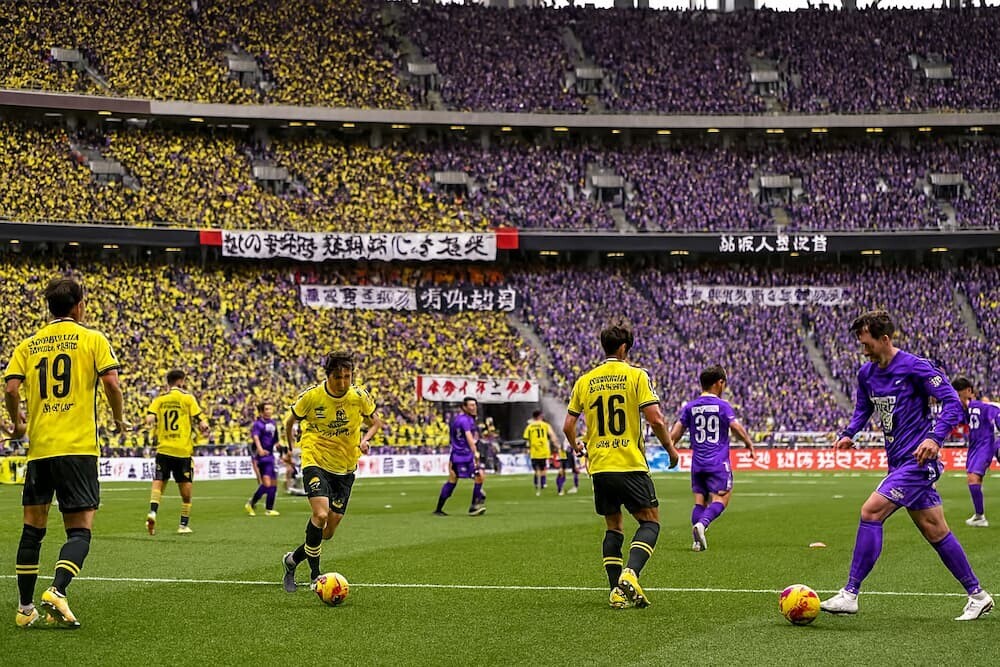 国立競技場のピッチでウォーミングアップする柏レイソルと広島の選手たち、両チームのサポーターで埋め尽くされた満員のスタンド、日本人選手と日本語の応援幕を生成AIで作成したリアルなイメージ