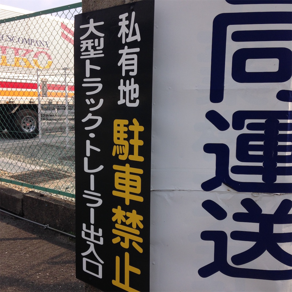 黒地に白で「私有地」、黄色で「駐車禁止」、白で「大型トラック・トレーラー出入り口」と書かれた、縦書きの看板。右に「なんとか同運送」という白い看板があり、どうやら運送会社の敷地のようだ。