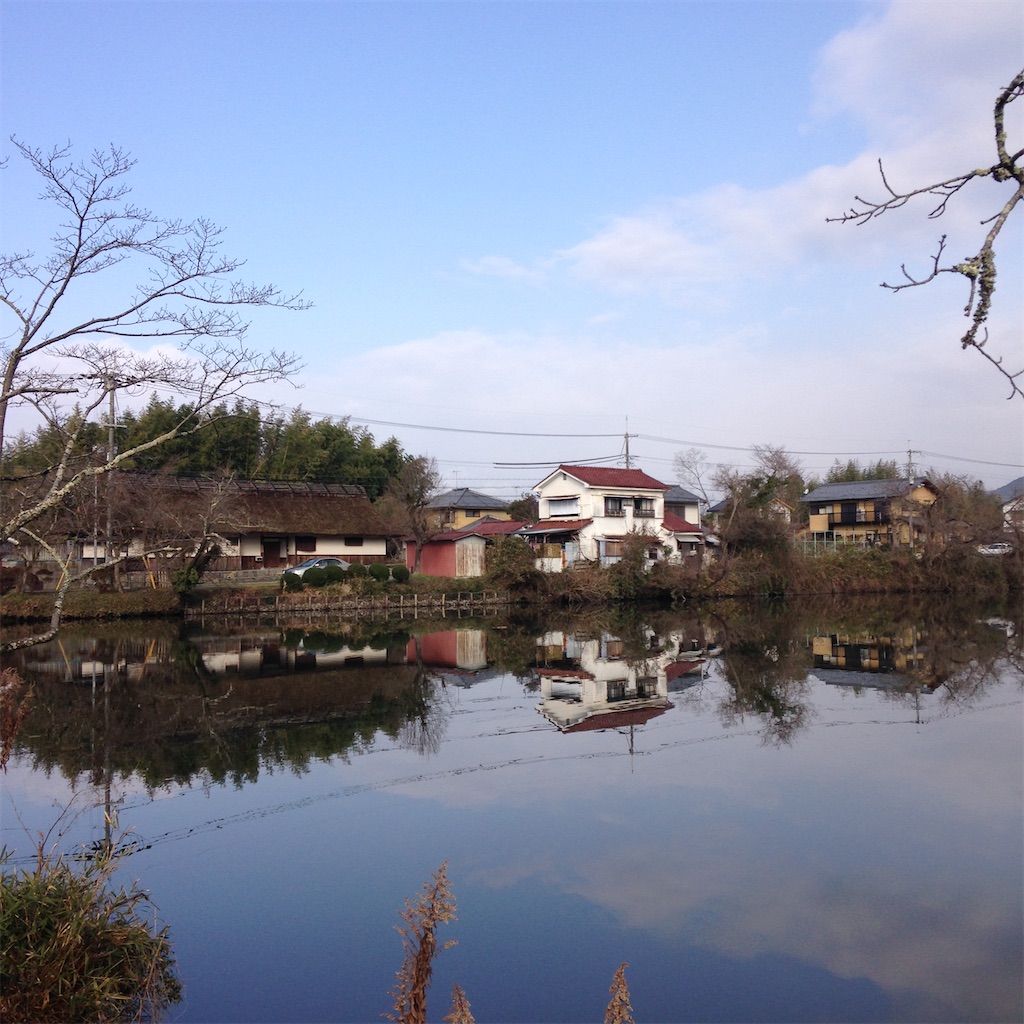 手前に池、水面に向こうの家々や木が反射している。向こうには茅葺き屋根の家と、赤い屋根に白い壁のかわいい家。空は穏やかに晴れている。