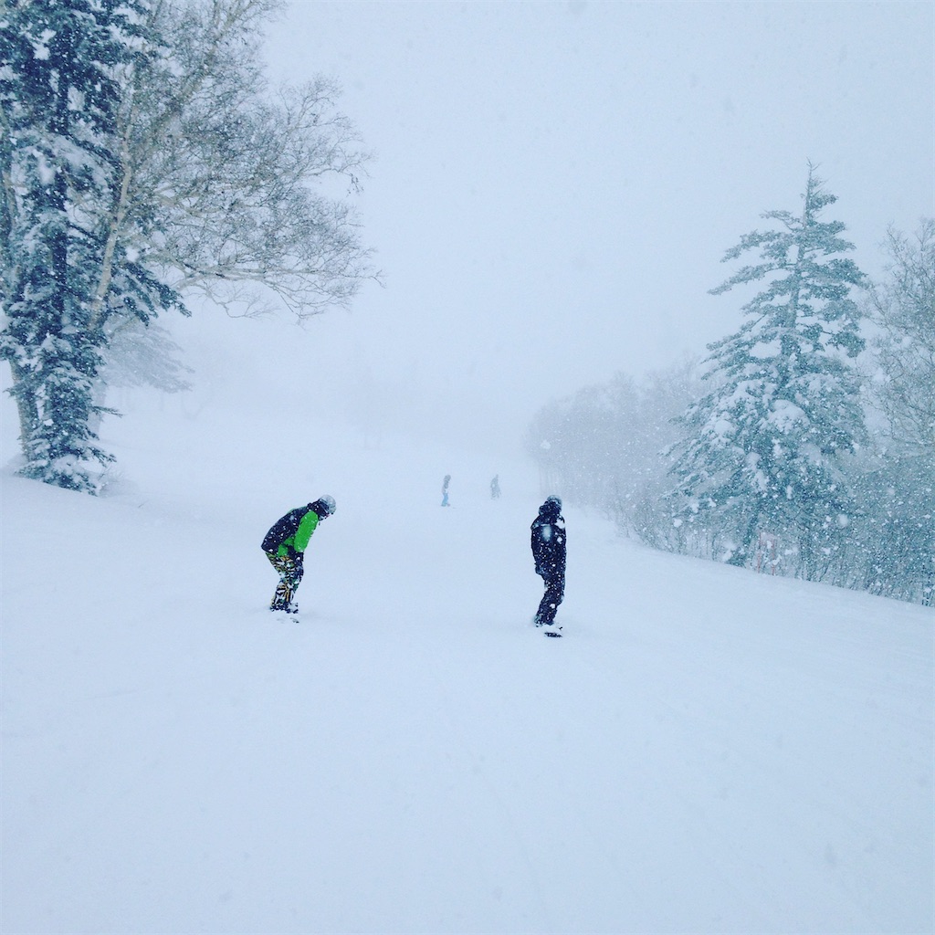 雪が舞うなか、山のなかのちょっと平らな部分で、向こうに二人のスノーボーダーがいる。左右には雪が積もった木々。