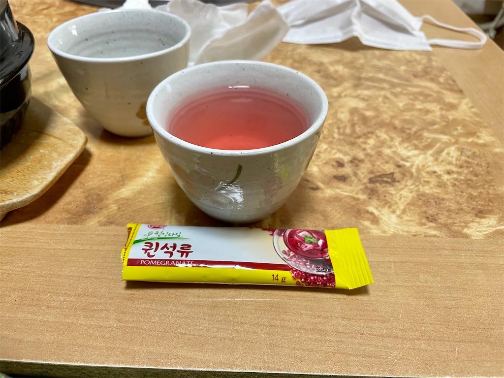 ピンク色のお茶っぽい飲み物が湯呑みに入っている写真。湯呑みの手前に「퀸석류」と書かれたスティック袋がある。