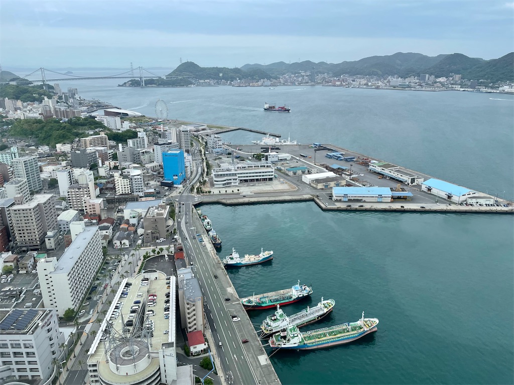 海峡ゆめタワーから下を見下ろした写真。手前に立体駐車場、湾岸に船が3隻係留されている。その奥に小さめの船が何隻か。その奥は小さな半島が突き出ていて、消防署や倉庫、コンテナが積んである場所。その奥に観覧車、ぬめっとした形の低い建物、ずっと奥には関門橋。