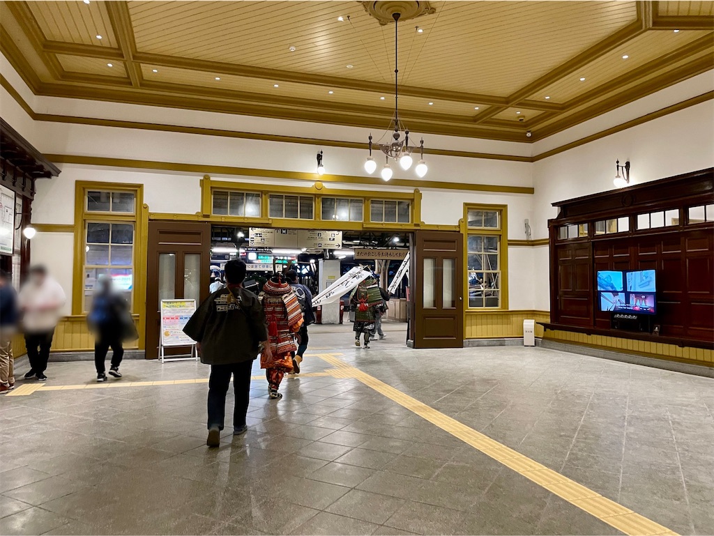 門司港駅の玄関に入った写真。照明が吊り下げられていて、白い壁に黄土色の窓枠や赤い引き戸がある、レトロなデザイン。武装した武士たちとお付きのスタッフが駅へ入っていく。