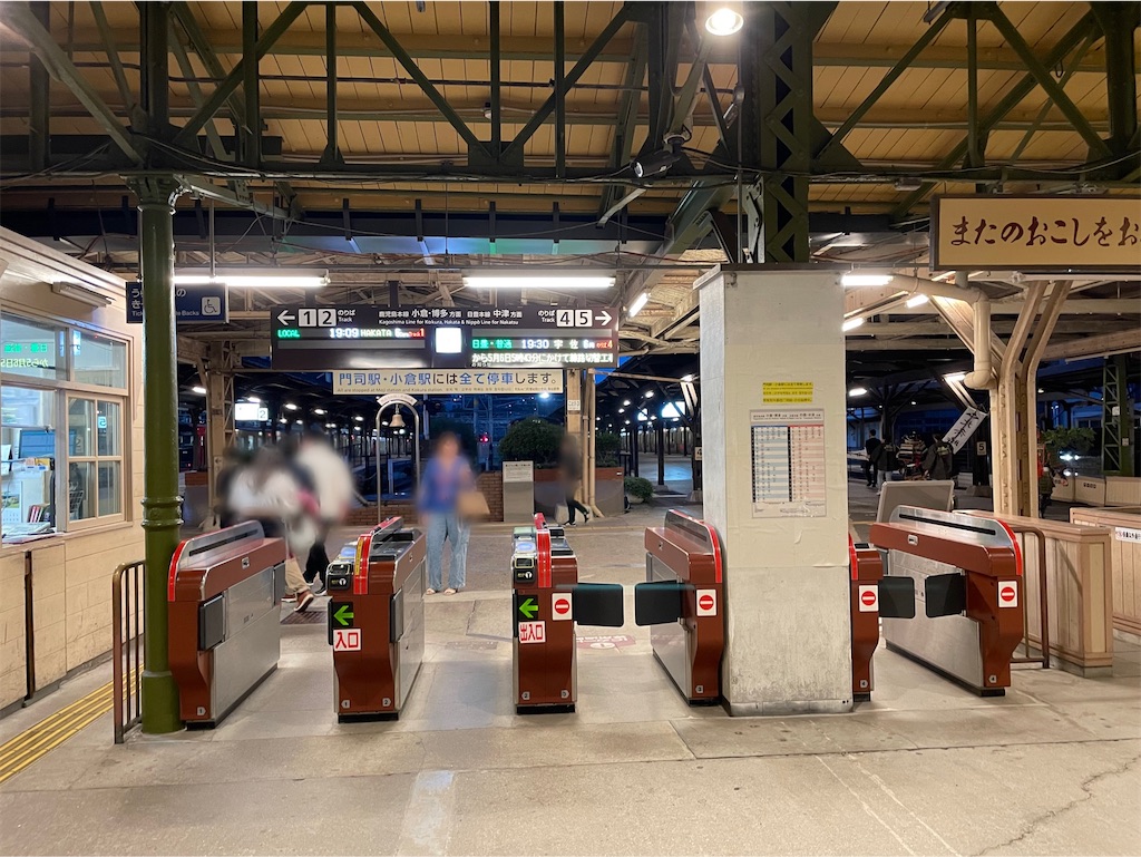 門司港駅の改札の写真。自動改札が4つ、電光掲示板に博多方面と中津方面が表示されていて、下に「門司駅・小倉駅には全て停車します」という横断幕が下がっている。数人の人影。