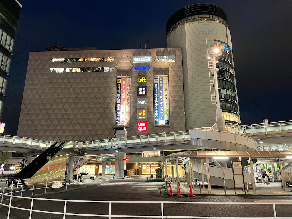 駅前のロータリーの写真。大きいペデストリアンデッキの向こうに、ショッピングビルがある。LOFT、無印良品、ベスト電器、ユニクロなどの店名が壁に掲げられている。ロータリーには人も車もほとんどいない。