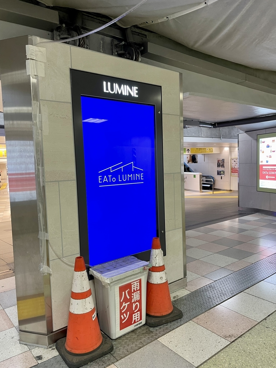 LUMINEのディスプレイ前に置かれた「雨漏り用バケツ」と貼られた角形ゴミ箱、ゴミ箱には天井に張られたシートから伸びるホースが接続されている
