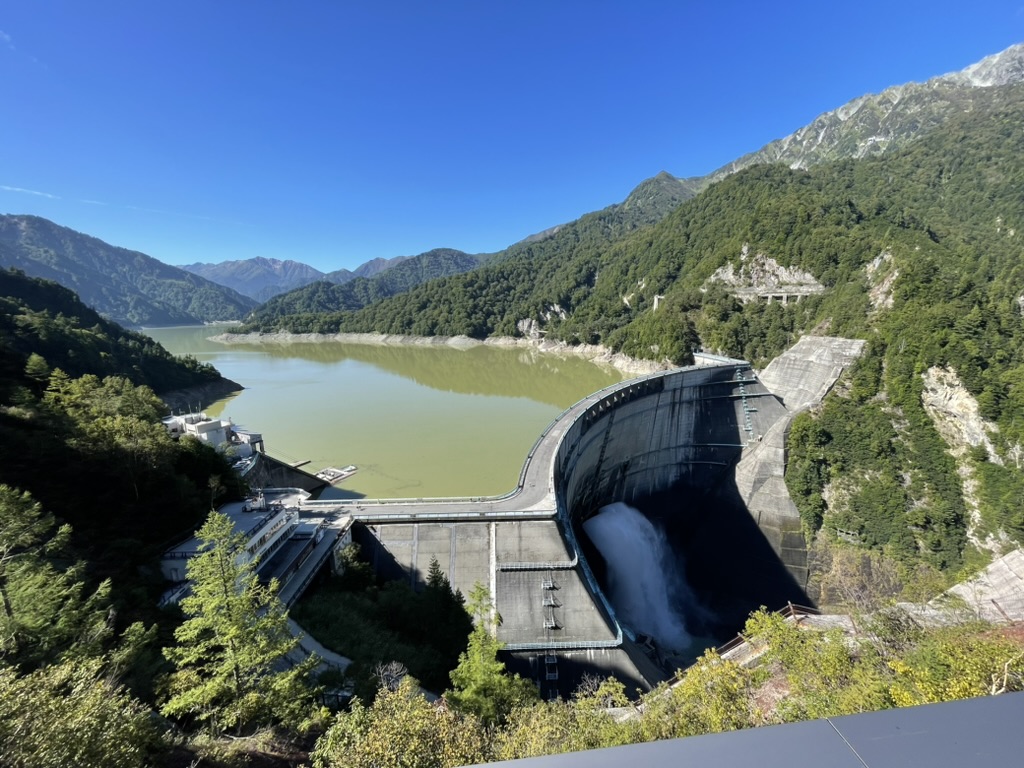 青く雲ひとつない空、山に囲まれて緑色の水をたたえたダム、中央にダムをせきとめている壁があり、壁の中央から水が勢いよく吹き出している
