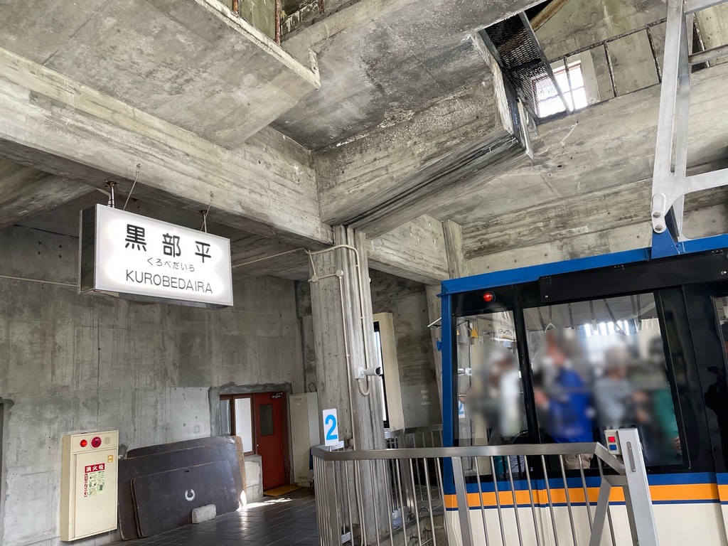 「黒部平」の駅名標、コンクリ打ちっぱなしの無骨な駅、人が満杯に乗ったゴンドラがとまっている