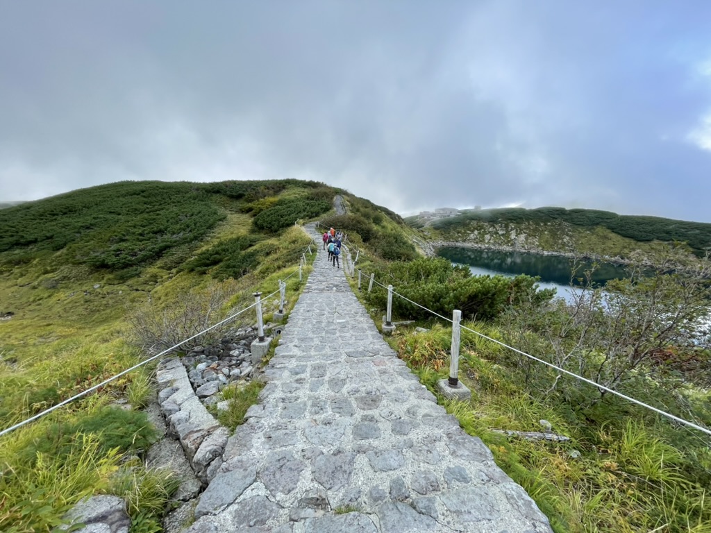 中央に石畳の道、左右に草や低い木、右下に鏡面のような池。空は曇りっていうか、霧がかっている。