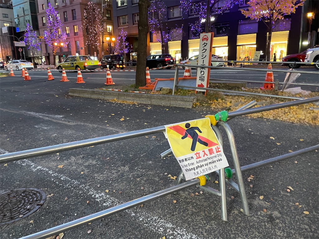 工事中の歩道を区切る柵に下げられた旗っぽいやつ、歩くピクトさん。背後に御堂筋、タクシー乗り場の看板。道の向こうは夕暮れで木にクリスマスの電飾が光っている。