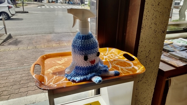 小布施駅の観光案内所入り口付近になる消毒液の写真。消毒液に毛糸でつくったかわいいカバーがかけてある。目と口がついていて、ほほえんでいる。