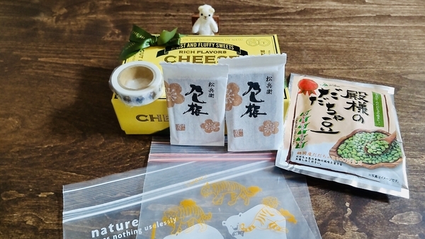 おみやげの写真。だだ茶豆のスナック菓子やのし梅、焼き菓子などのおみやげを小さなくまのぬいぐるみが見ている。