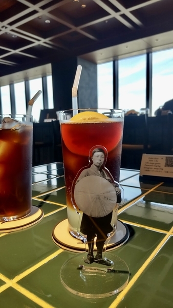 カフェの写真。テーブルにアイスコーヒーがのっていて、その手前に正岡子規のアクリルスタンドが立っている。