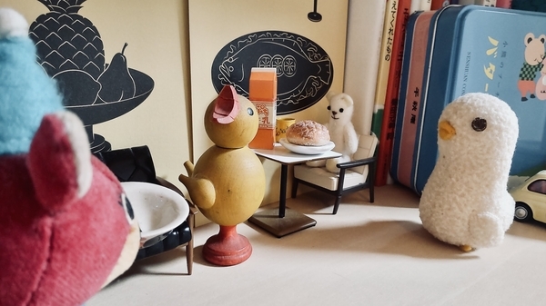 本棚の一隅を撮った写真。小さなぬいぐるみや木彫りの鳥がテーブルを囲んでいる。テーブルには牛乳やパンが出ている。