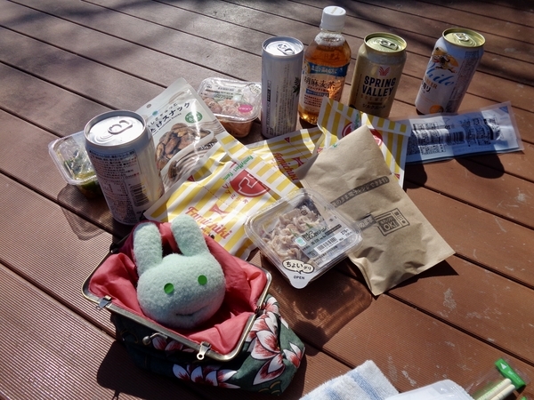お昼ごはんの写真。コンビニで買ったお酒やおつまみが公園のテーブルにのっている。ふかふか手まくらもある。