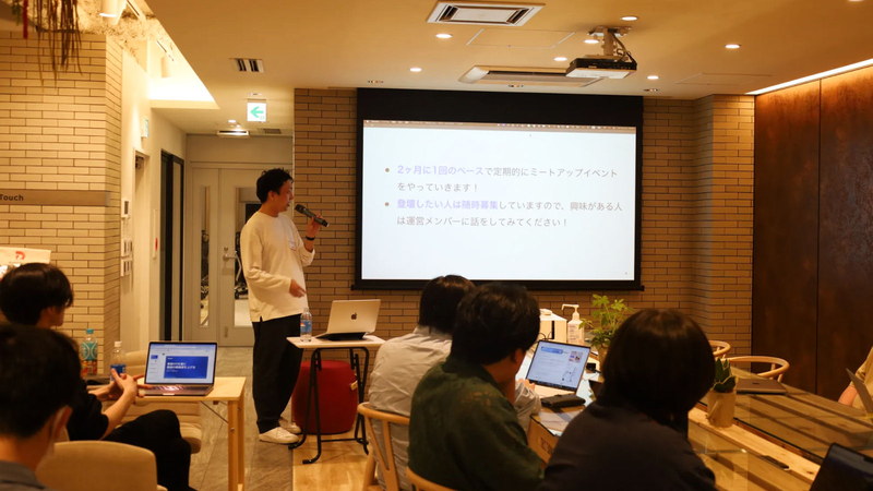 マイクを持つ登壇者がスライドを使ってイベントの案内をする様子。
