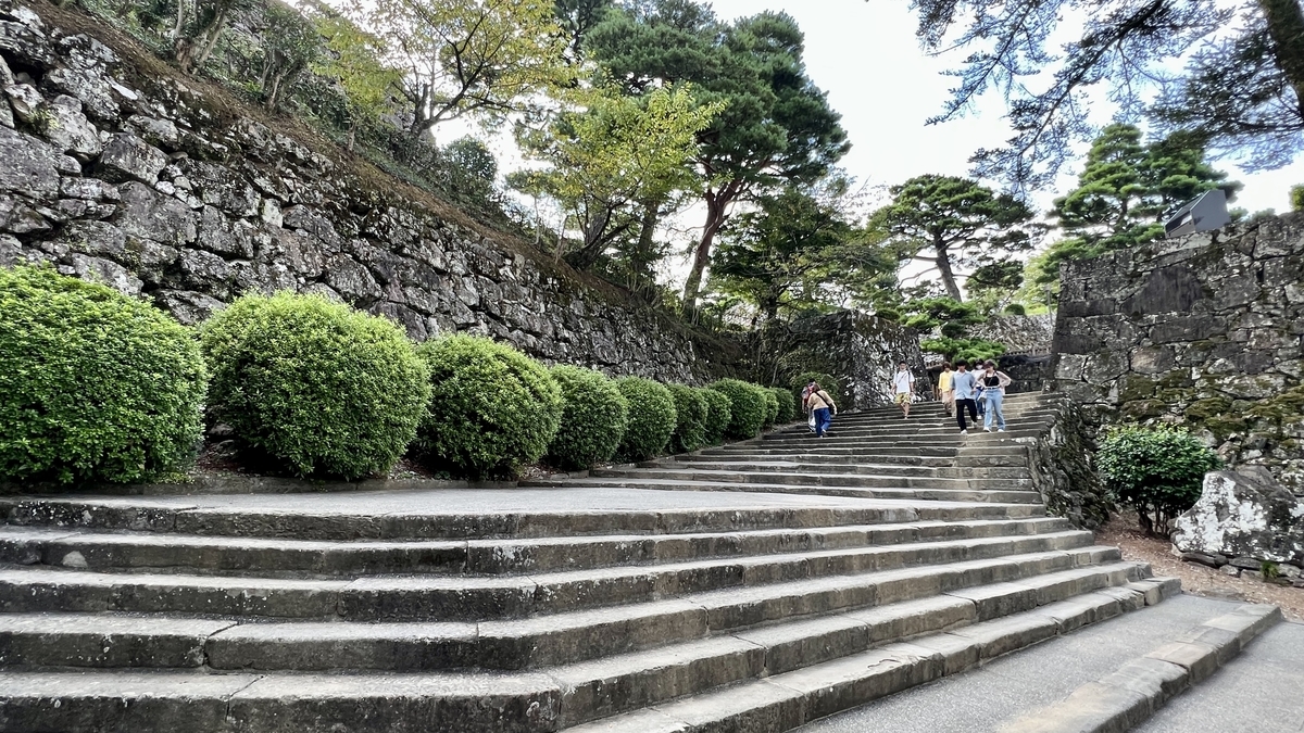石垣に囲まれた緩やかな石段を登る人々。左手には整えられた植栽が並ぶ