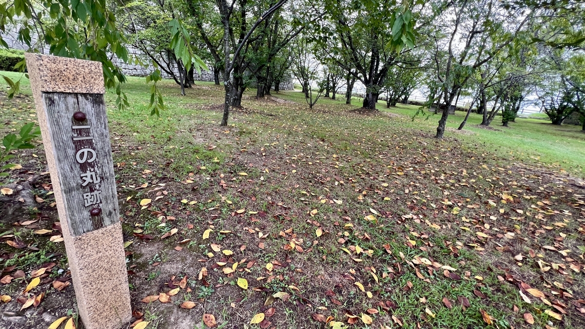 「二の丸井戸」の案内標柱と木立に囲まれた地面。井戸の入口に続く場所