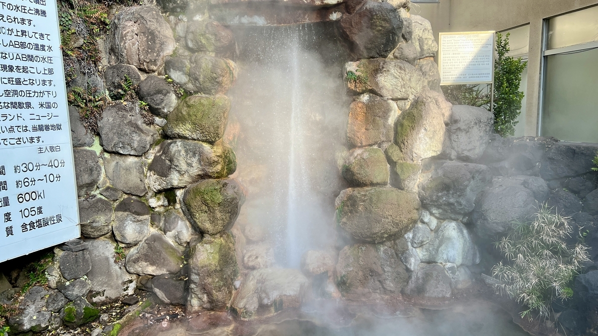 岩組みの間欠泉から白い噴気が上がり水しぶきに虹が出る様子