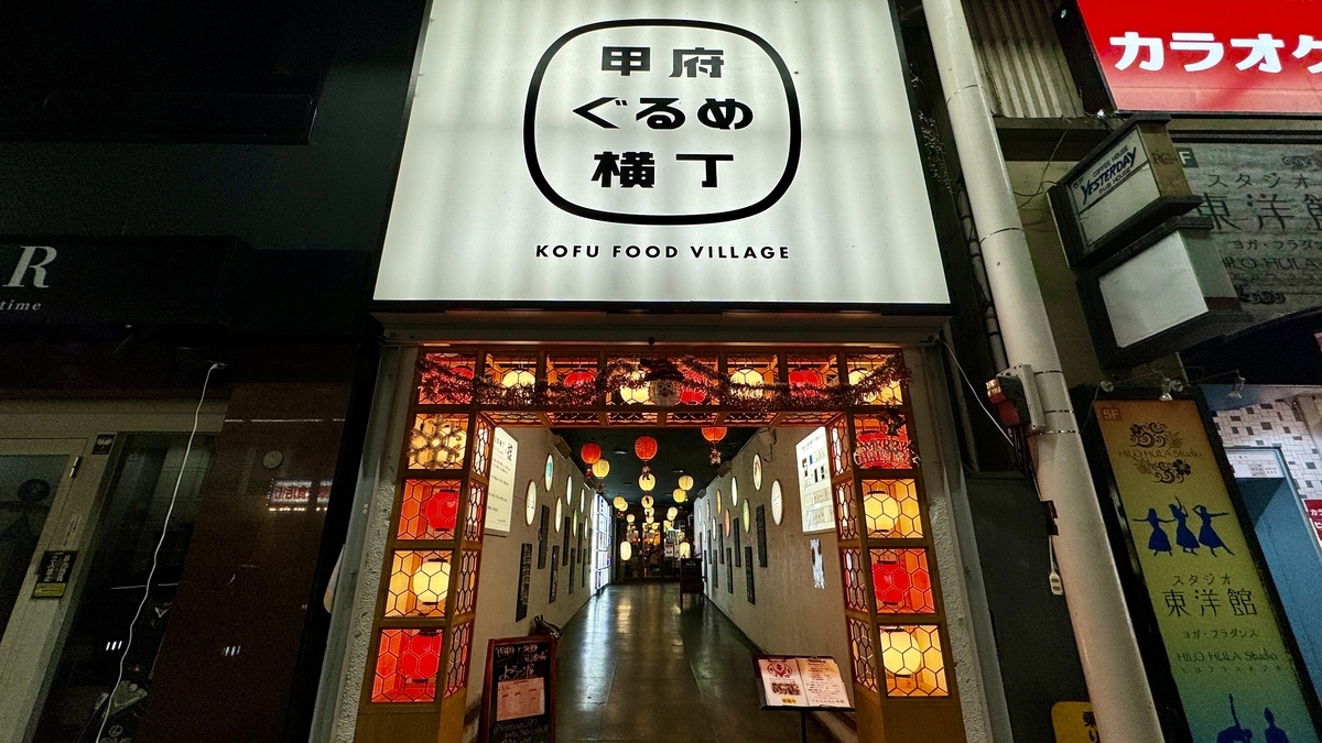 甲府ぐるめ横丁の入口で白い看板の下に赤い提灯が並ぶ通路