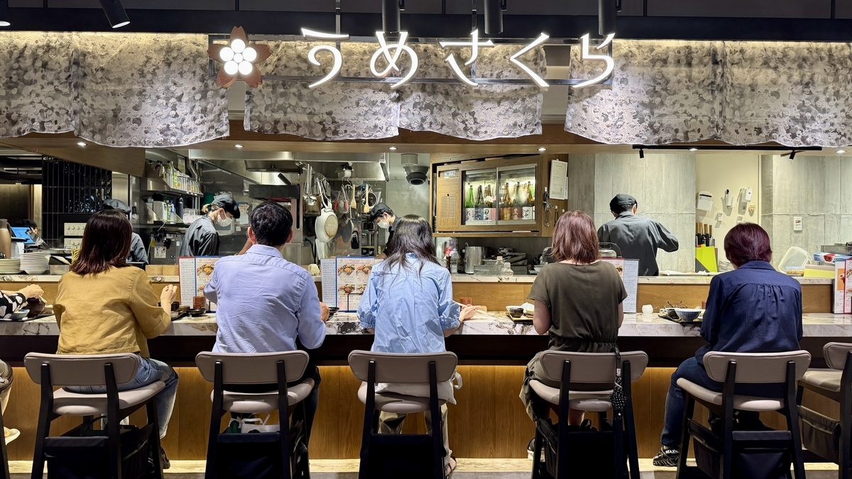 阪神バル横丁内のうめさくら店舗。木の温もりあるカウンターで客が並び、和食とお酒を楽しんでいる。