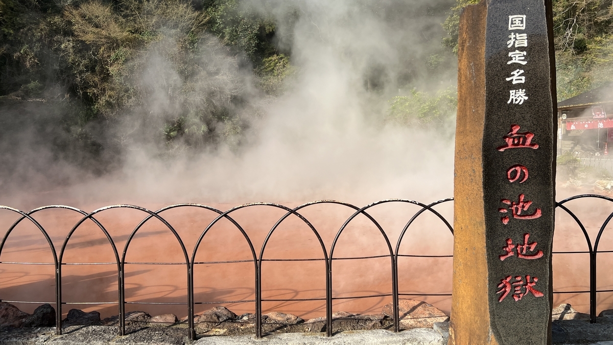 血の池地獄の赤い温泉池から湯気が立ち上り手前に血の池地獄の石柱が立つ様子
