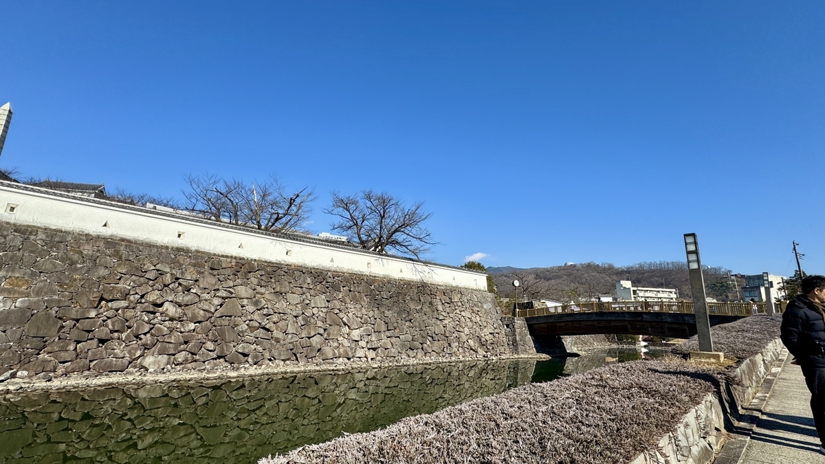 甲府城の石垣と堀の水面が続く風景 青空の下に橋と遊歩道が伸びる昼の景色