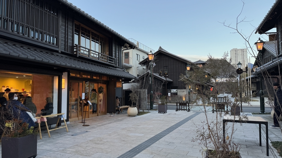 夕暮れの広場に灯る街灯と町屋の明かり カフェの窓辺で過ごす人々とベンチのある景色