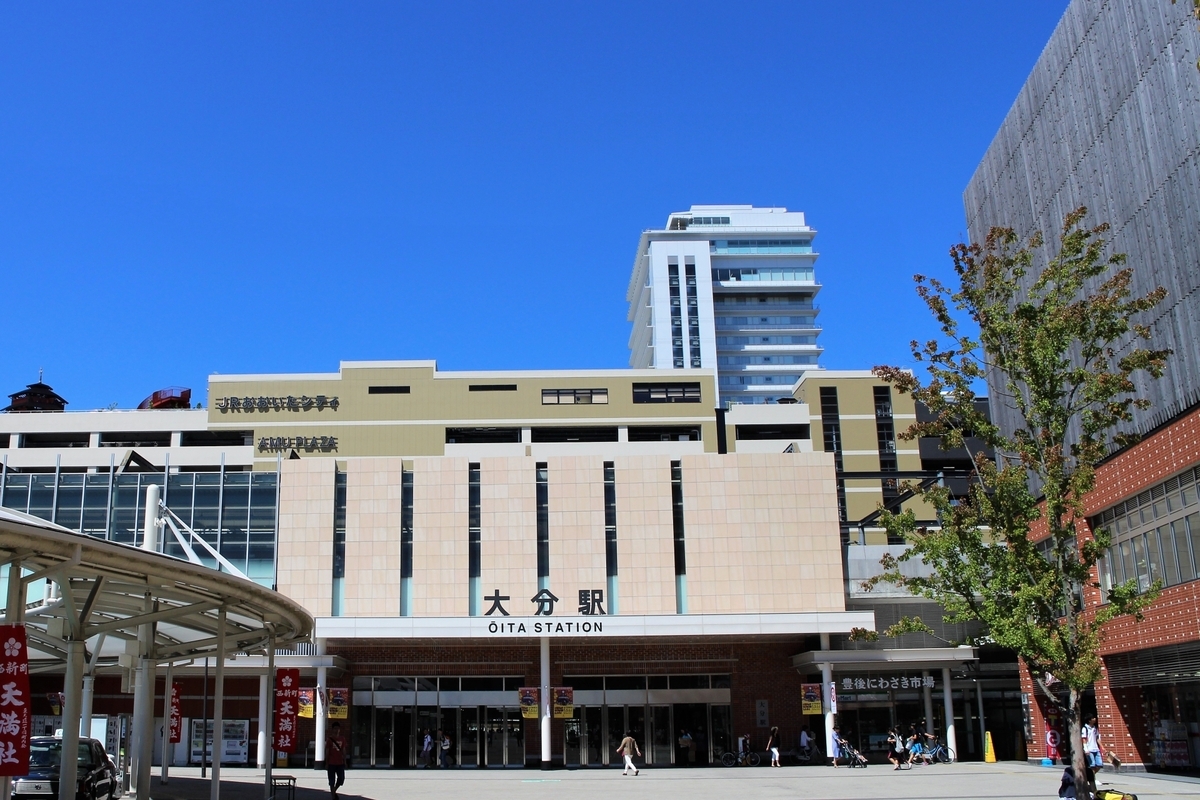 青空の下に大分駅の駅名看板が見える駅ビル正面と人の行き交う駅前広場