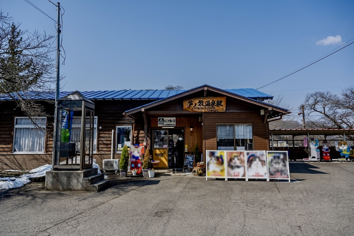 青い屋根の木造駅舎に芦ノ牧温泉駅の看板が掛かる駅前と猫の写真パネル