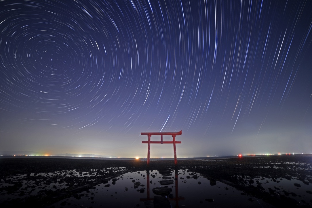 干潟に立つ赤い鳥居が水面に映り星の軌跡が描かれる夜空