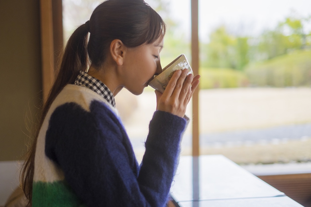 窓辺のテーブルで湯呑みを両手で持って抹茶を飲む女性の横顔