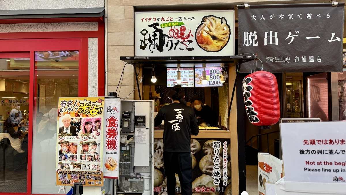 踊りだこ道頓堀店の店頭で大きな看板と券売機が並び店員が焼き場でたこ焼きを焼く様子