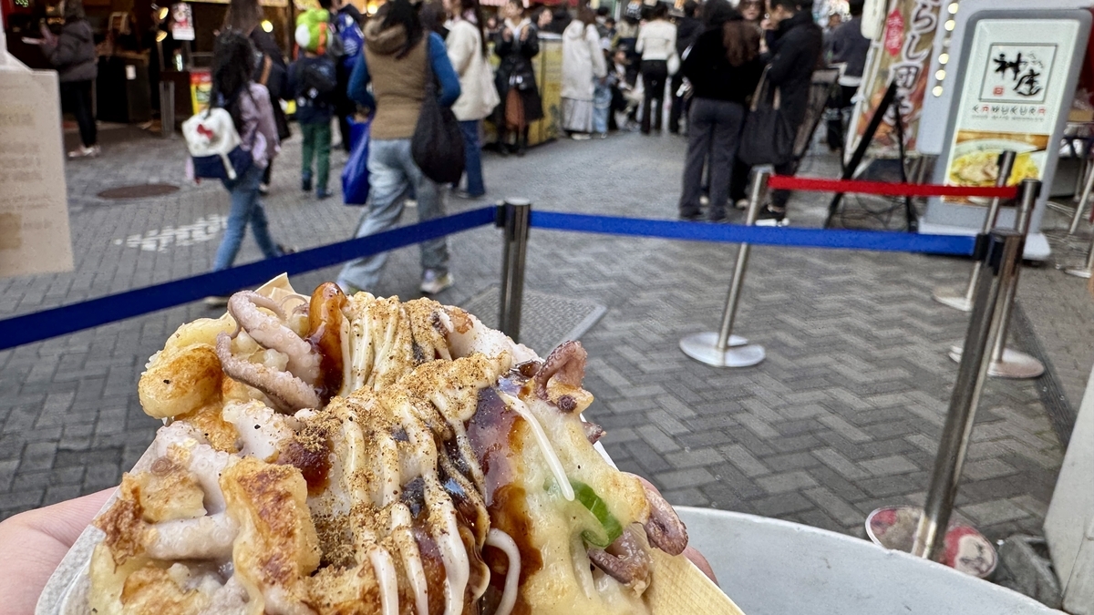 踊りだこのたこ焼きを手に持ち道頓堀の行列とロープ柵が見える風景