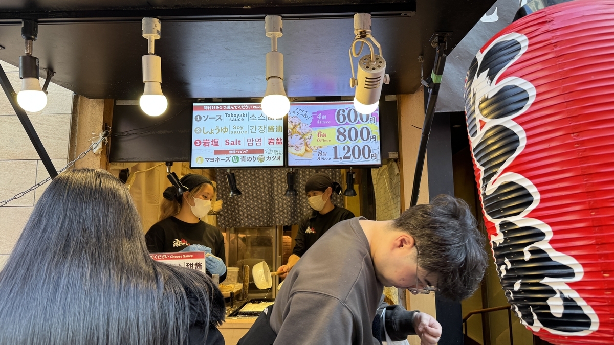 踊りだこの店頭でスタッフが鉄板でたこ焼きを焼き上げる様子と味付けと価格の看板
