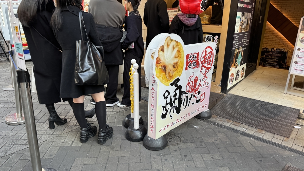 踊りだこの店頭に置かれた丸ごとイイダコたこ焼きの看板と並ぶ客の列