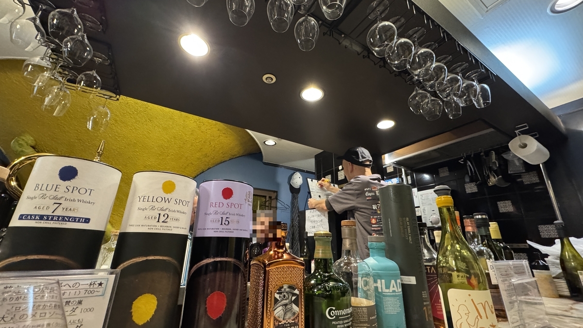 天井から吊るワイングラスとカウンターに並ぶウイスキーやジンのボトル 奥で店主が作業する店内