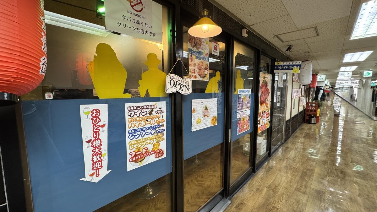 青い腰壁と黄色い人物シルエットが並ぶnighthawksの外観 おひとり様歓迎の札が掛かる入口